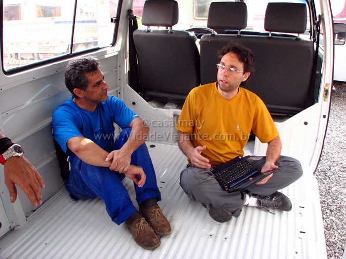 a man with a laptop sits on the ground with another person in a van