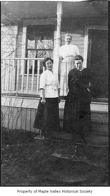 two woman stand in front of a house while another woman stands on the porch