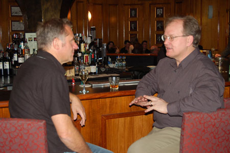 two men sitting at a bar in the evening talking