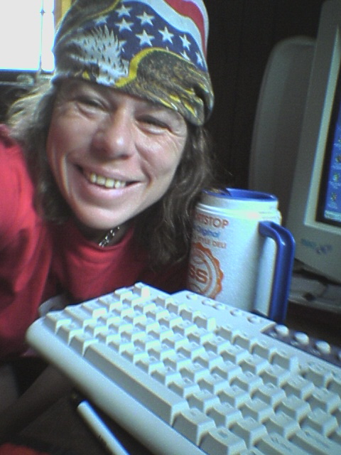a man wearing a beanie smiles next to a computer keyboard and drink