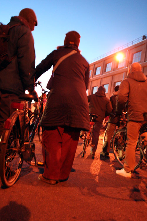 several people stand around with bicycles and buildings in the background
