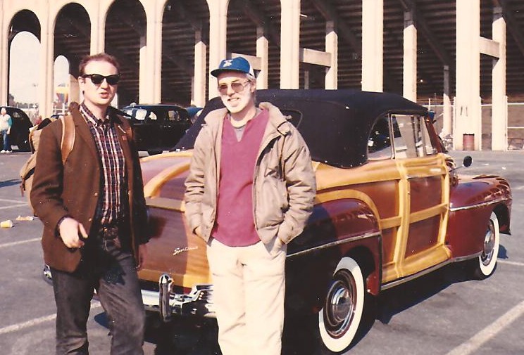 two men standing next to an old car in a parking lot