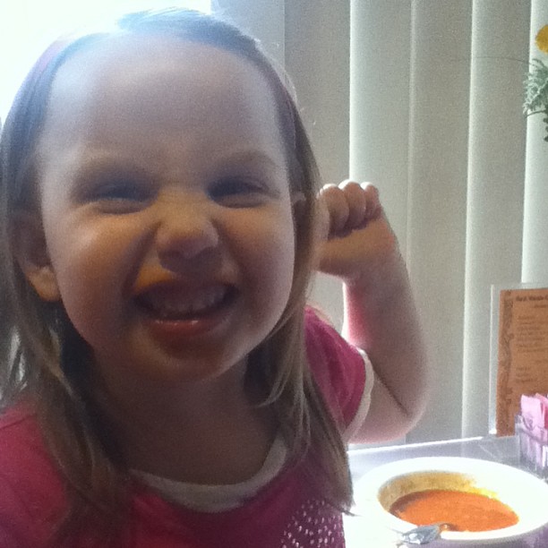 a little girl smiles while eating a bowl of soup