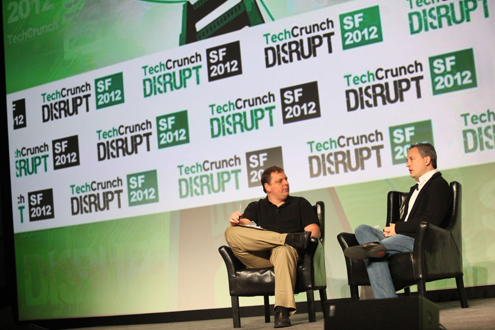 two men sitting on chairs in front of a green wall