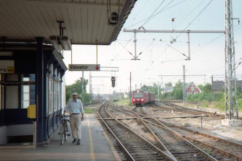 a man in a white shirt walks along a railway platform with a bicycle and train tracks