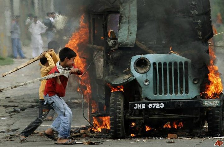 two children walking by a burning jeep with people nearby