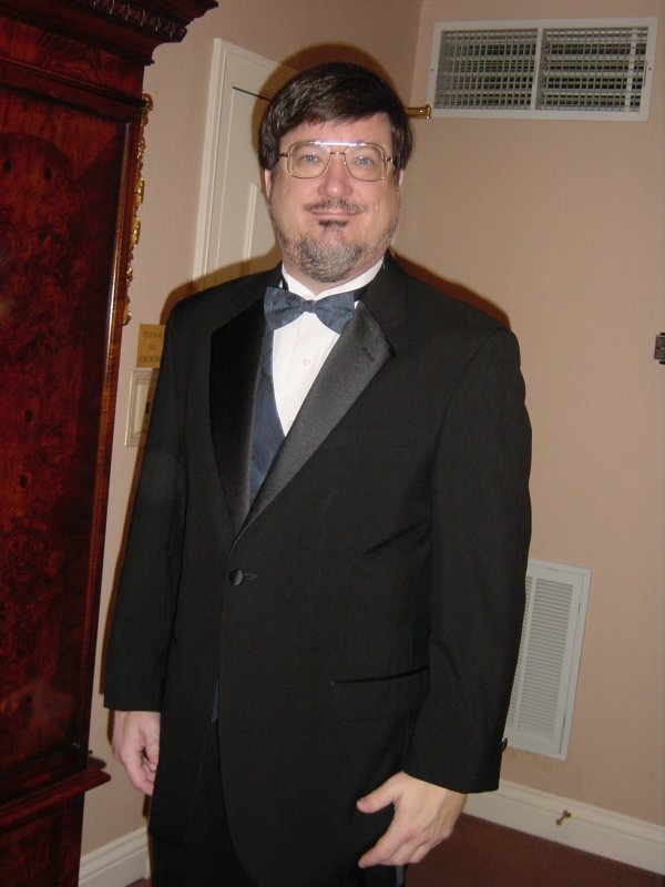 man wearing glasses and a black tuxedo posing in front of a grandfather clock