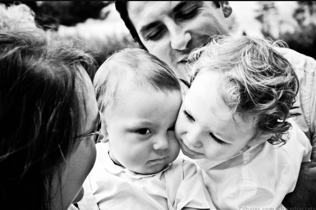 two little children kiss their mom's lips while she is surrounded by their parents