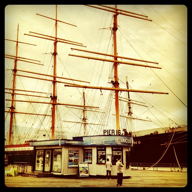 two people stand at a small shop on a wooden dock in front of a ship