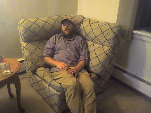 a man in glasses and a hat is sitting on a couch with beer in the corner