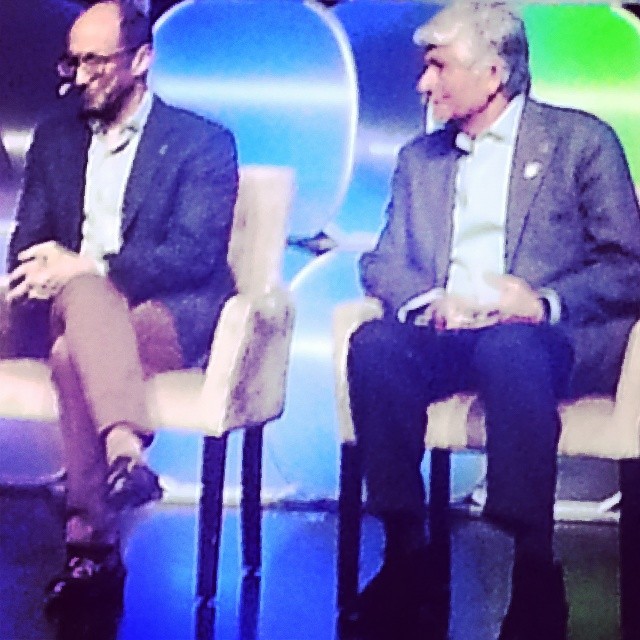 two men sitting down in chairs talking while wearing suits