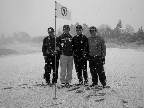 four men wearing winter jackets in snow near a flag pole