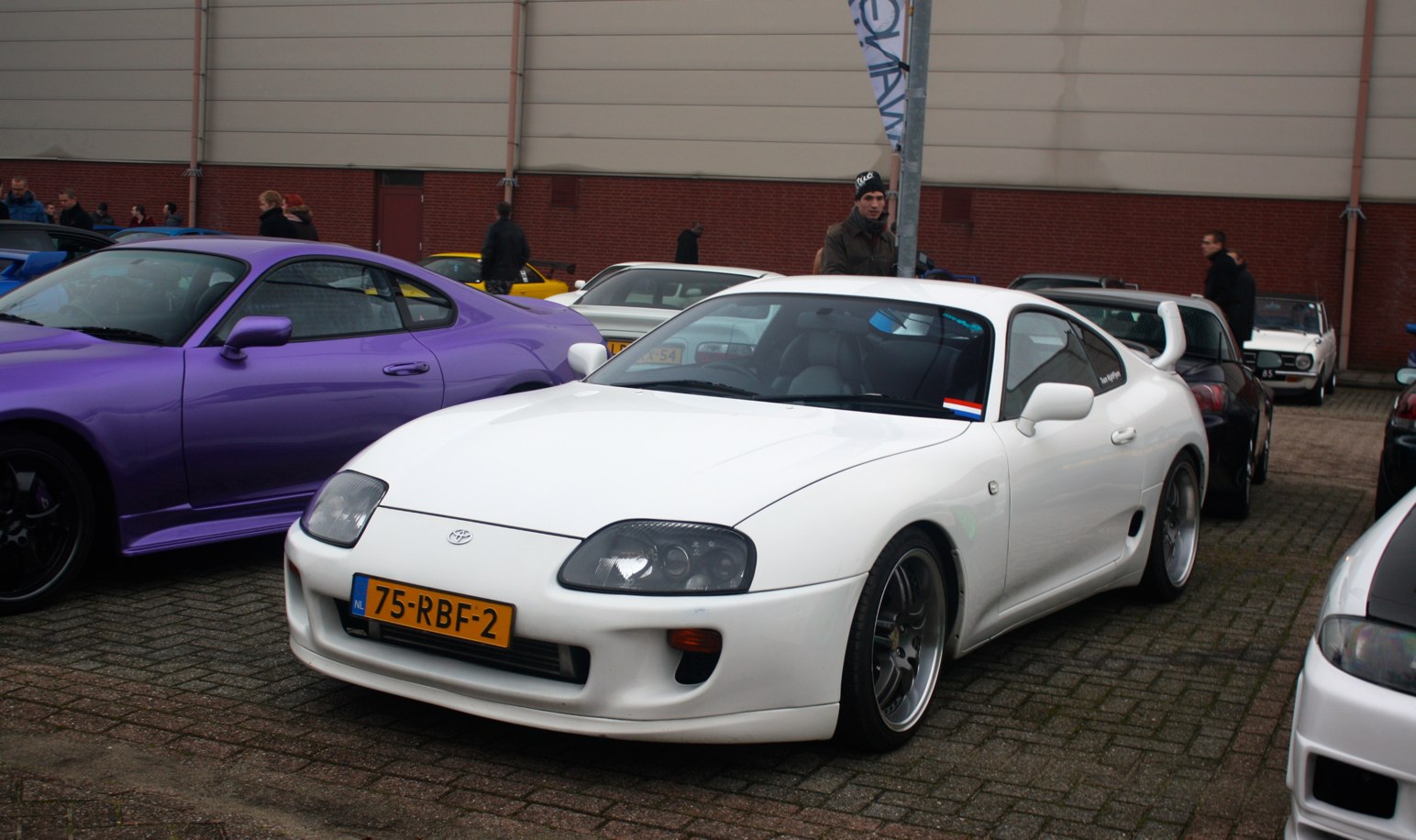 three parked sports cars in a lot and some people standing on the side walk