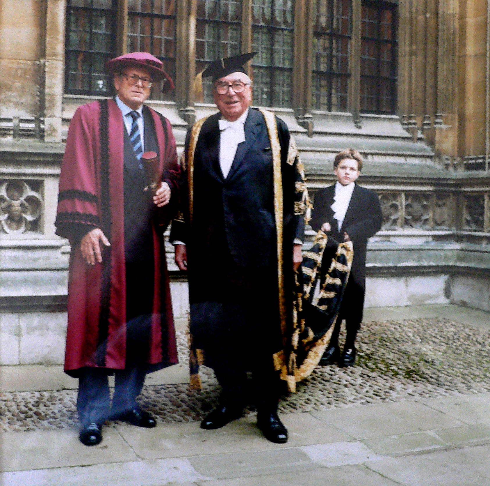 two men in gowns and one in a graduation cap are standing on a stone walkway in front of a large building