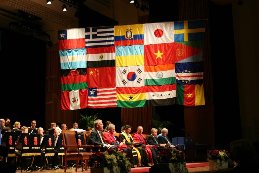 an assembly hall with people sitting on stage and flags in the background