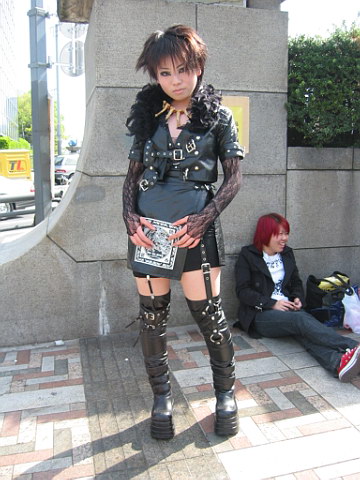 a young woman dressed up in punk clothes and knee high boots, is standing near a stone wall