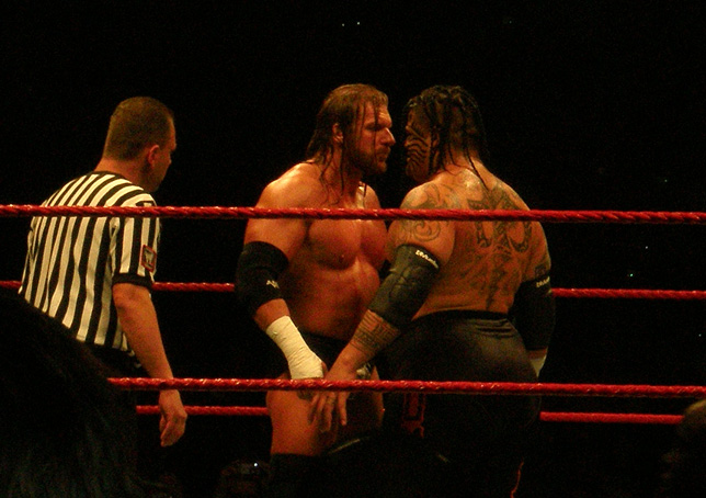 men standing in a ring with the referee, two with wrestling gear, and one with a  top