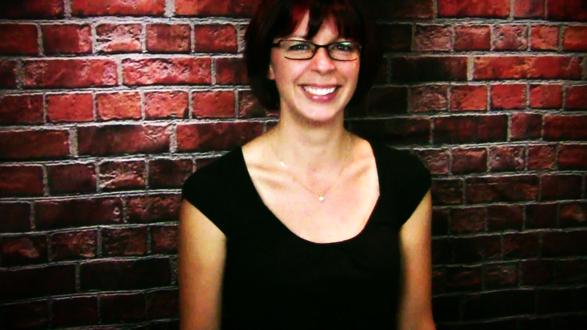 a woman with glasses smiles next to a brick wall