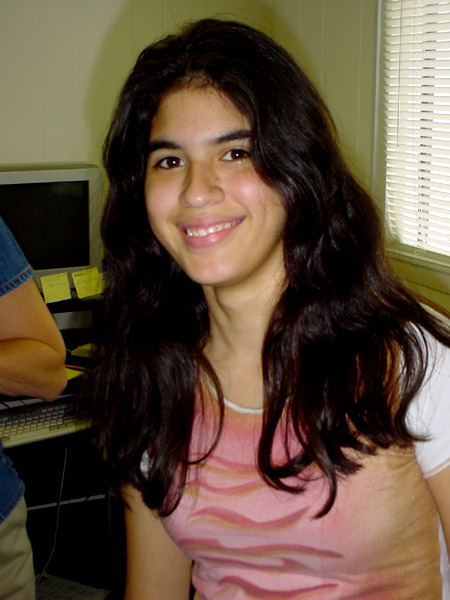 a girl with long hair and a pink shirt smiles in front of a computer