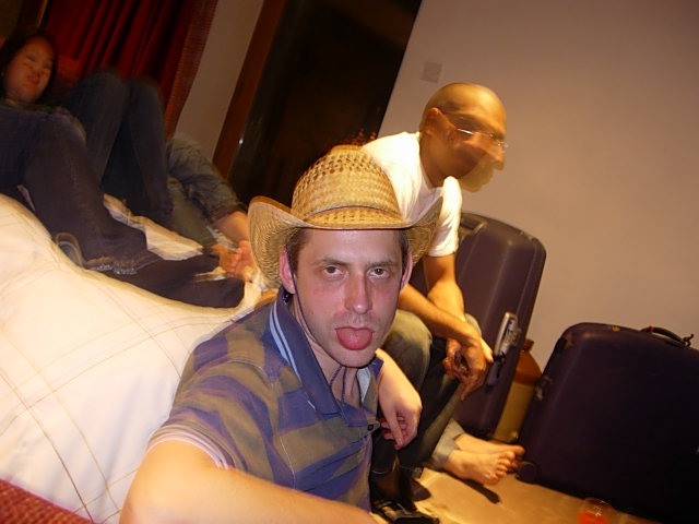 a man sitting on a bed with a hat on his head and people standing near by