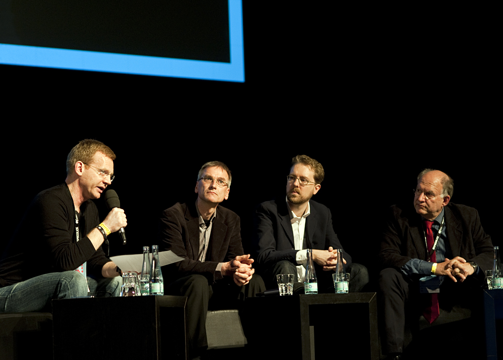 four people sitting on black chairs at a panel in a dark room