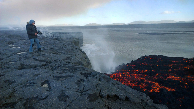 a man on a rock looks at the lava and smoke