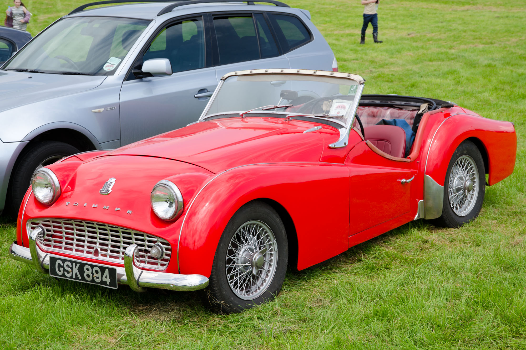 an old red sports car on grass with a couple of people looking at it