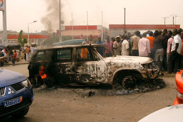 people standing in front of a burnt car with the fire burning