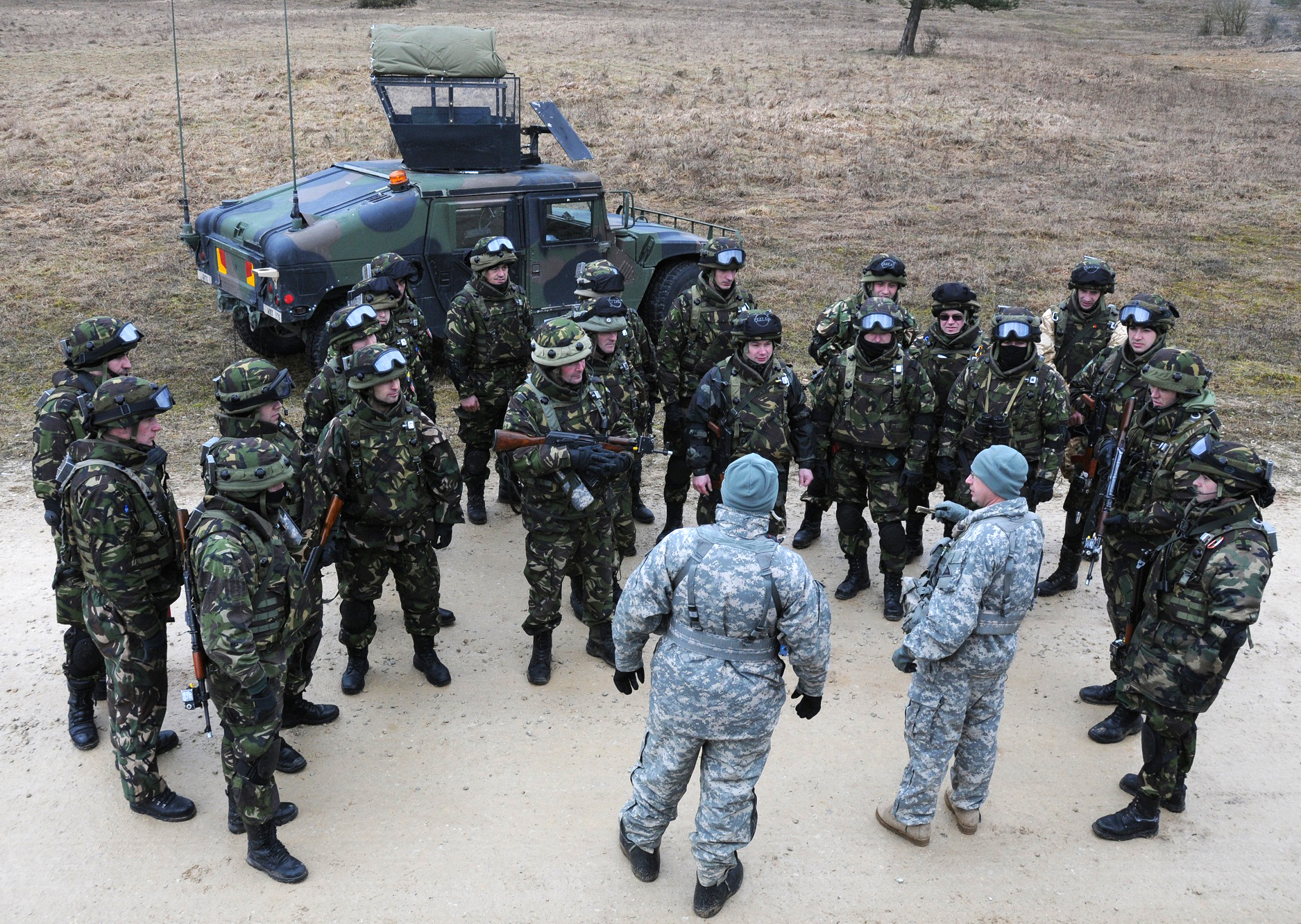 military personnel in uniform standing in the dirt with an armored vehicle