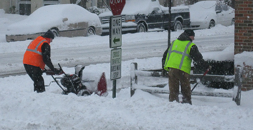 two workers shovel snow from a sidewalk