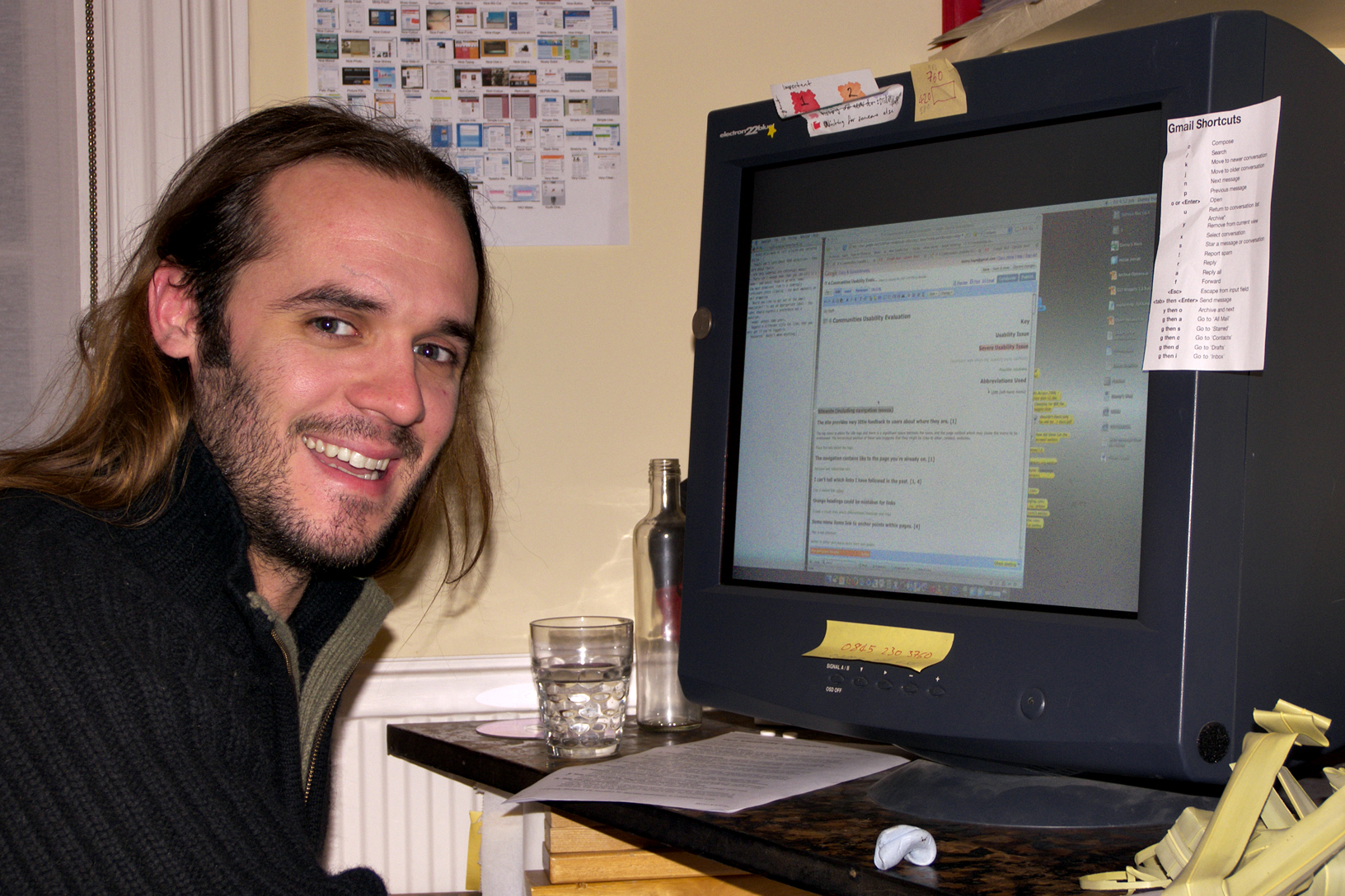 a smiling man with long hair sitting next to a computer screen