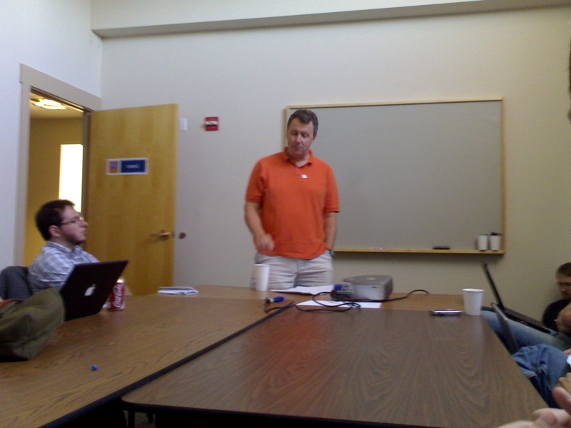 a man is standing and gesturing while sitting at a desk with a laptop