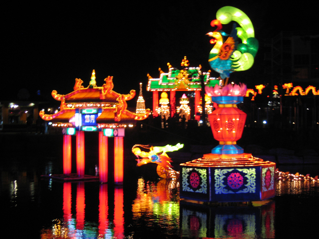 a lighted water fountain next to the water in front of colorful buildings