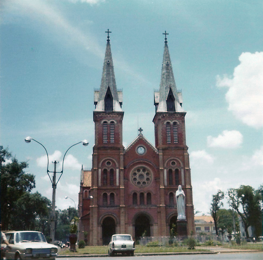 cars drive by an old cathedral under cloudy skies