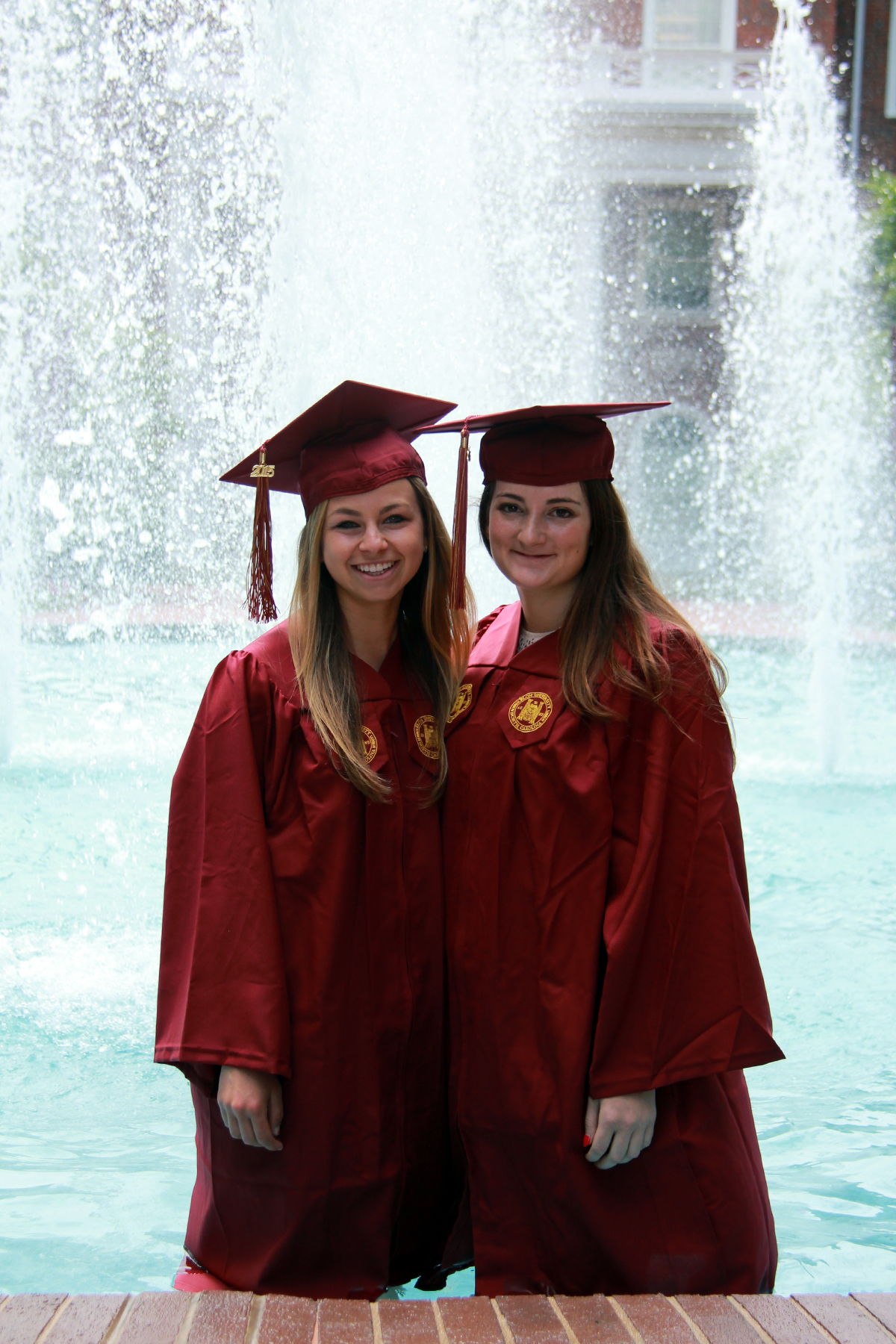 two graduates standing by the fountain in red caps and gowns