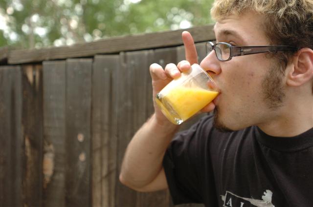 a man drinking an orange juice in front of a wooden fence