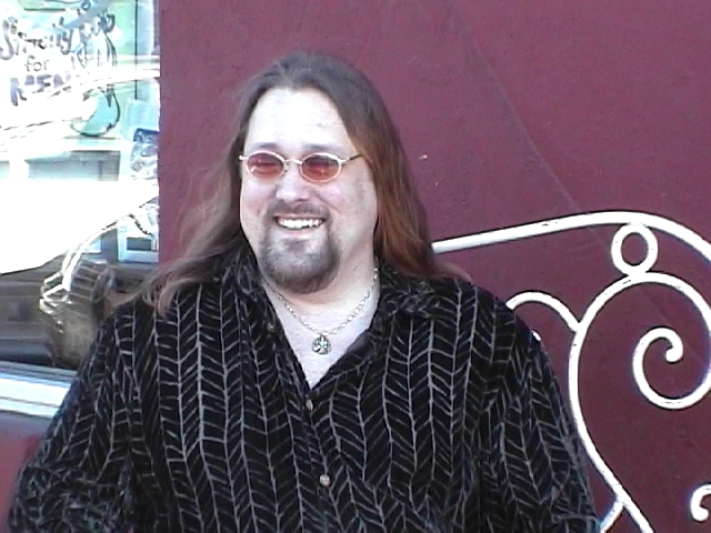 a man with long hair and glasses wearing an ornamentally designed shirt and smiling