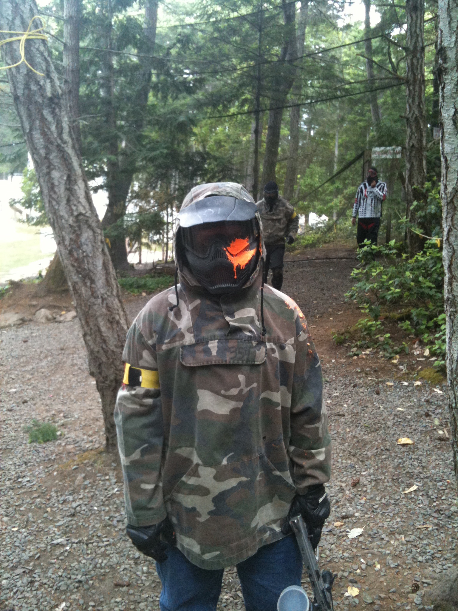 a man with a paintball helmet, hood up and standing in the woods