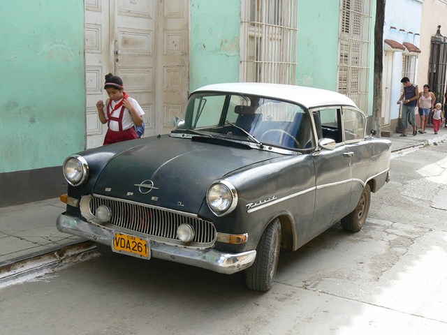 an old fashion car parked on the street, with people walking in front