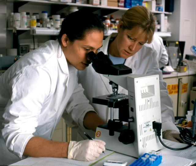two men wearing gloves are looking at an electronic device in a lab