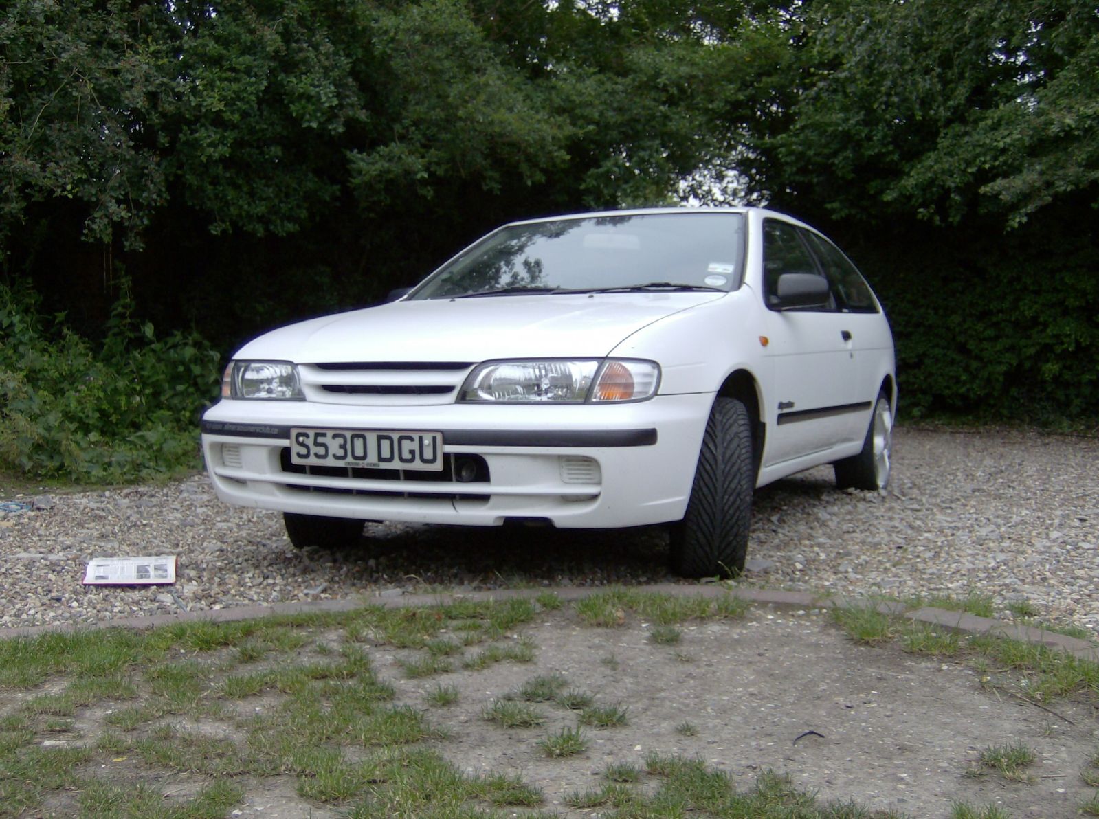 an older white car sits on a gravel area surrounded by trees