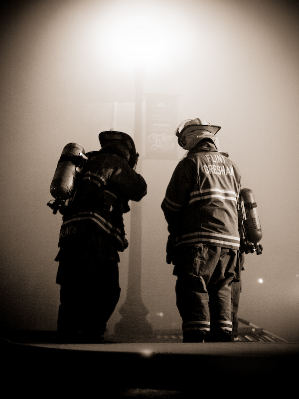 two fire fighters on the side of the street looking at soing