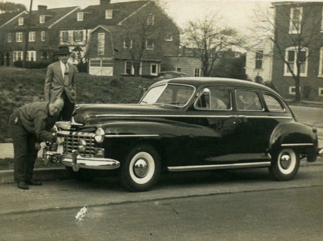 two men working on an old car with a motorcycle parked next to it