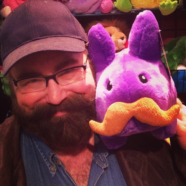 a bearded man is holding up a stuffed purple elephant with an orange nose