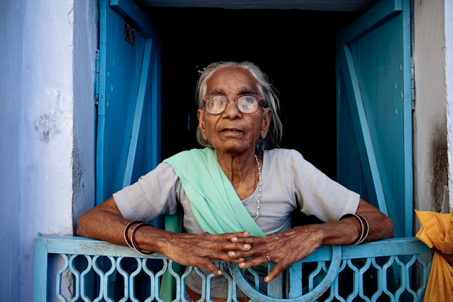 an elderly woman in eyeglasses and a green scarf standing outside of her window