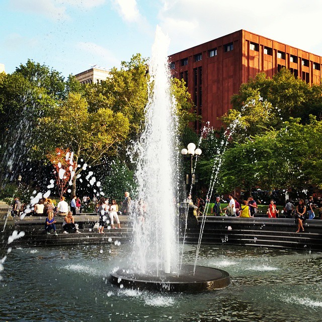 a large fountain of water in front of trees and people walking around
