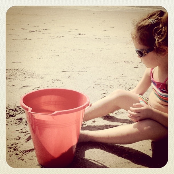a girl in a bathing suit and sunglasses playing with a sand pail on the beach