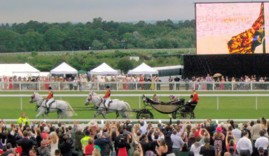 a group of people watching horses race with some riders in the cart