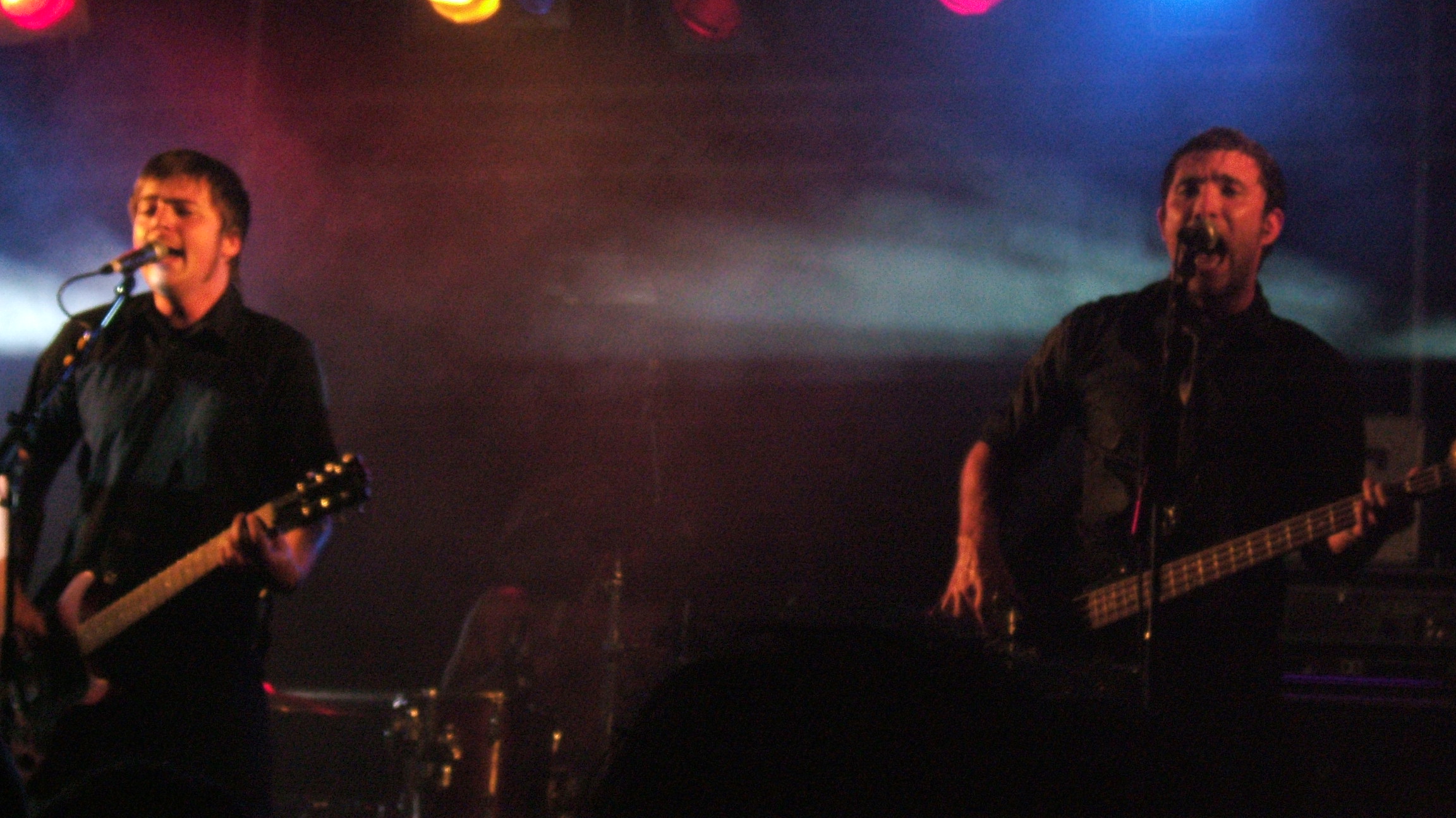 two men are playing guitar on stage with dark colors