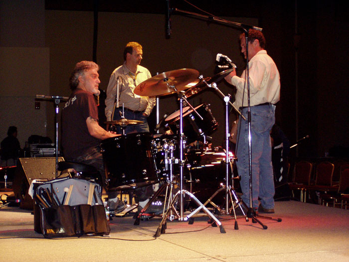 two men playing drums on a stage while another man listens to the speaker